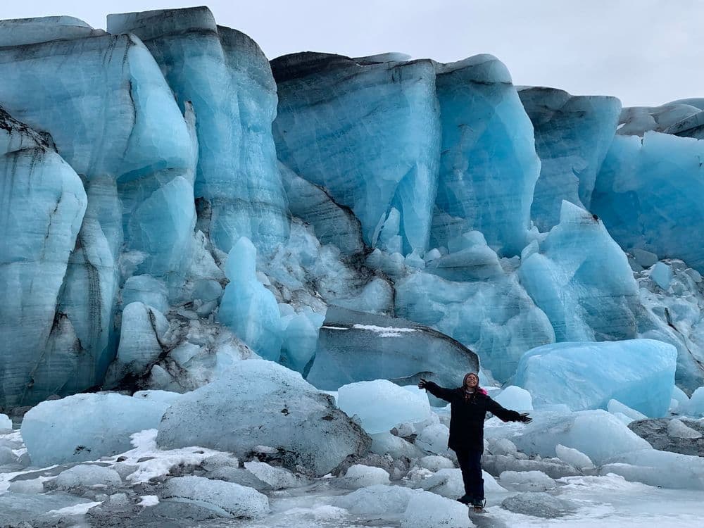 A person standing with arms outstretched at the foot of a towering blue glacier ice wall