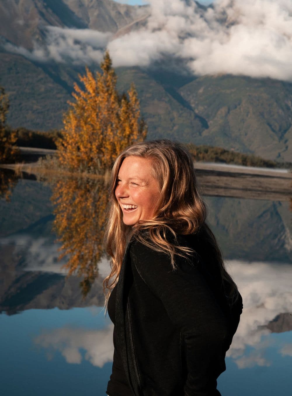 Kate Winter Evans laughing beside a still mountain lake in Alaska