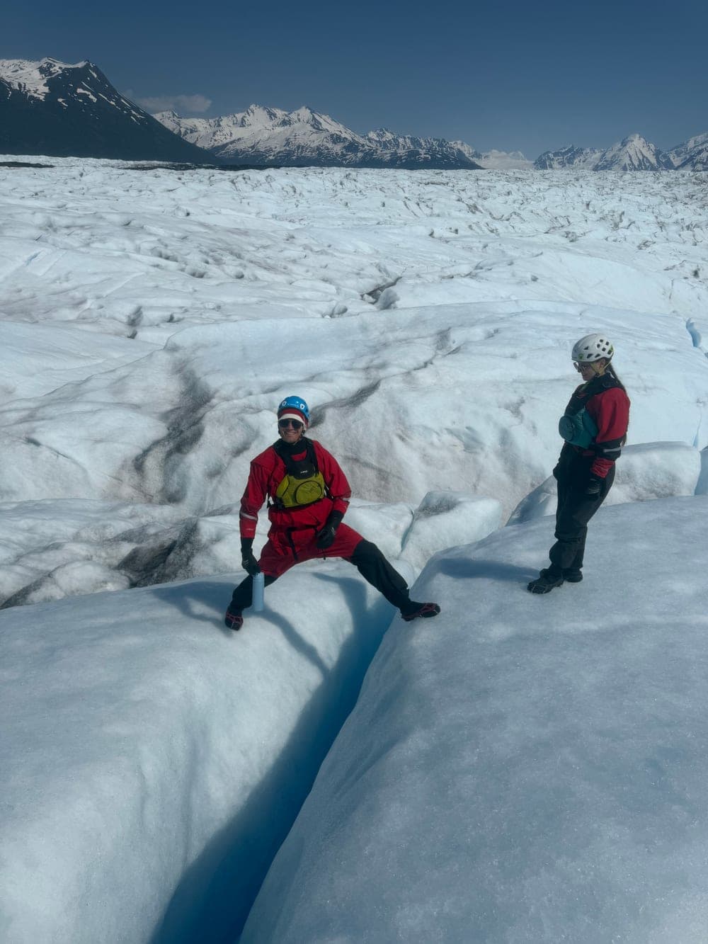 A guide in a red drysuit straddling a narrow crevasse and grinning, a second guide standing on the ice nearby