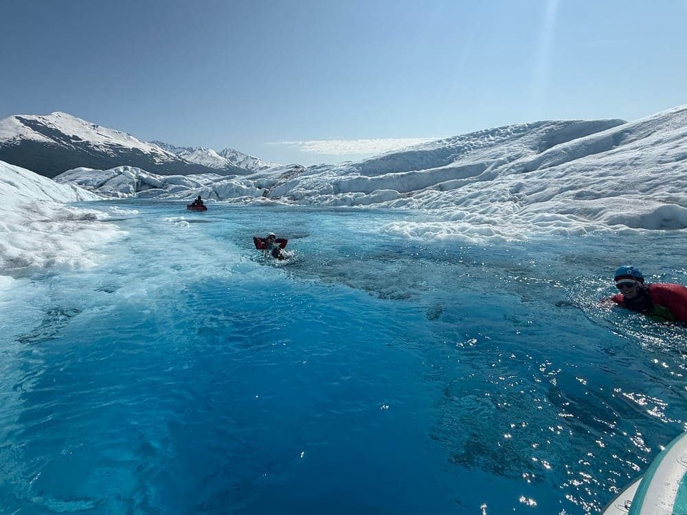 Three people in drysuits floating down a bright blue meltwater channel on a glacier, a red packraft ahead of them