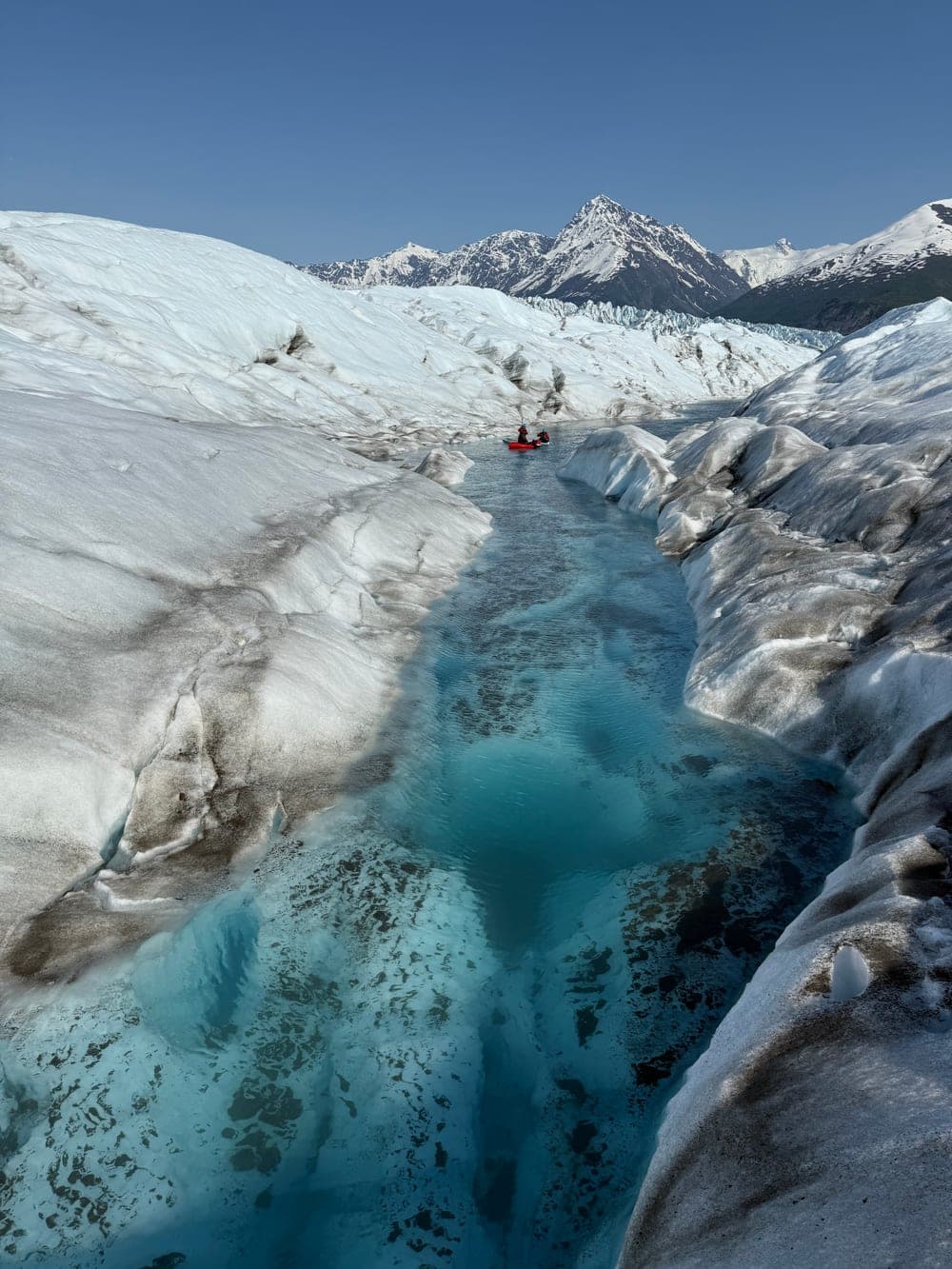 A red packraft carrying two paddlers along a deep meltwater channel cut into a glacier, a sharp snow covered peak beyond