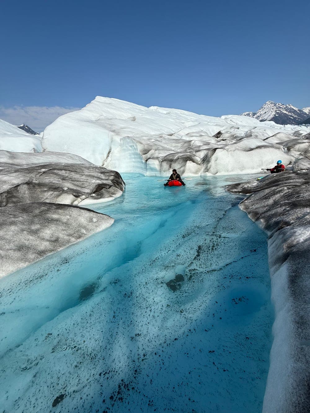 A paddler in a red packraft on turquoise glacial meltwater between banks of ice, a second paddler further along the channel