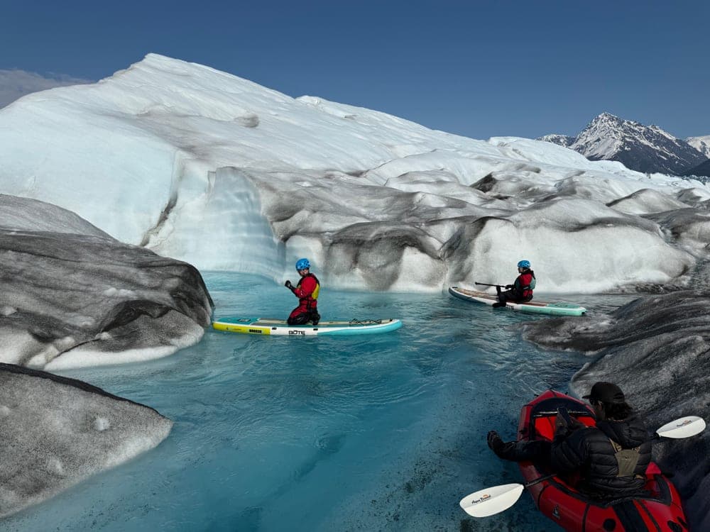 Two paddleboarders and a packrafter on a pool of pale blue meltwater beneath a wall of glacier ice