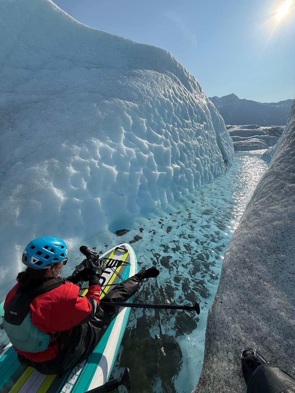 A paddler resting on a board beside a scalloped wall of glacier ice, looking down a narrow sunlit channel of meltwater