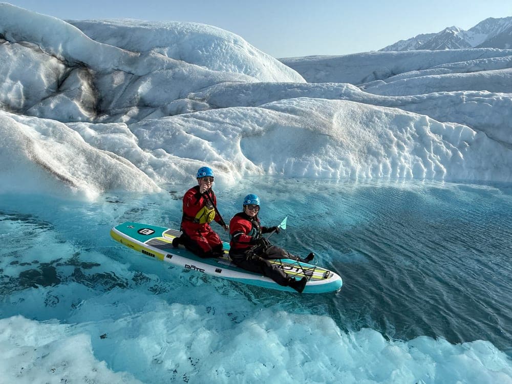 Two people sharing a paddleboard on a turquoise meltwater pool, with a bank of glacier ice and mountains behind them