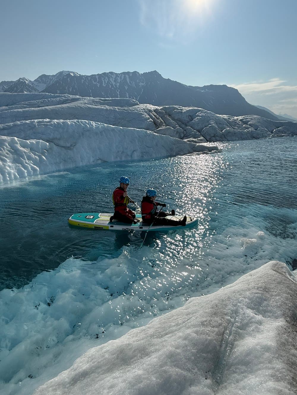Two paddlers sharing a board on open meltwater at the edge of a glacier, low sun glittering off the surface