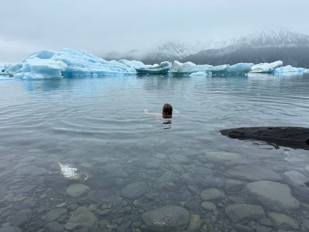 A swimmer out in a still lagoon among stranded icebergs, cloud hanging low over the mountains behind