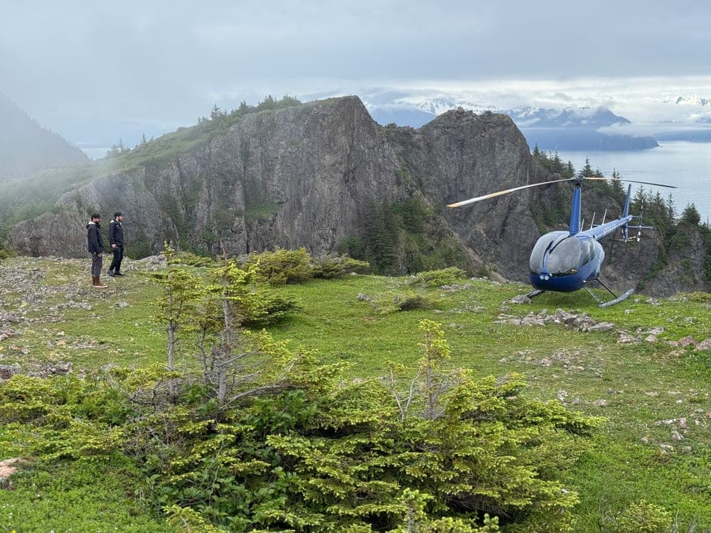 Two people standing beside a small blue helicopter on a green bluff above the sea, cloud over the mountains across the water
