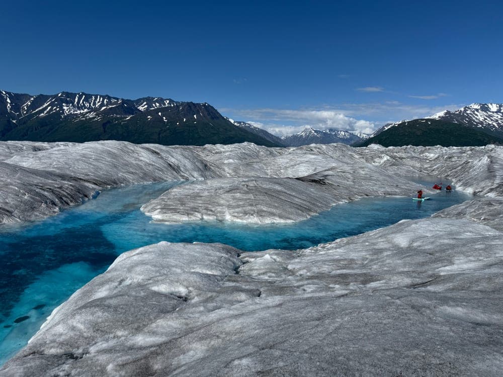 A blue meltwater channel winding across a wide grey glacier, with a paddleboarder and two packrafts small against the ice