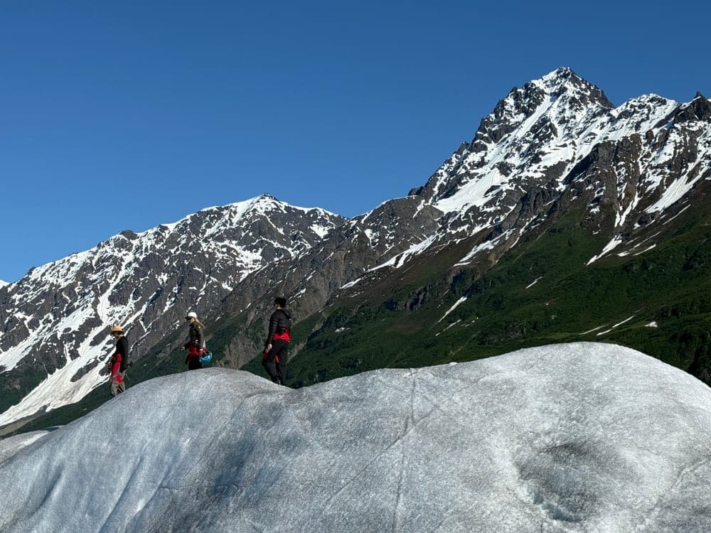 Three people walking along a rounded dome of glacier ice, a steep snow streaked peak rising behind them