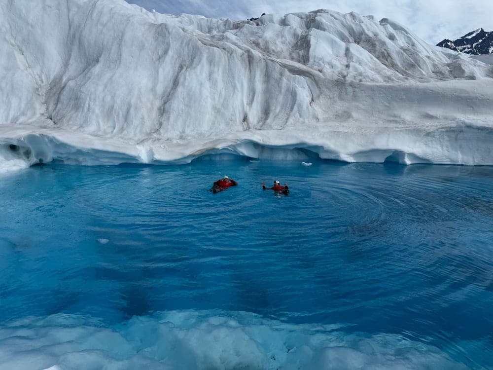 Two swimmers in red drysuits out in a wide blue meltwater pool, below a long wall of glacier ice
