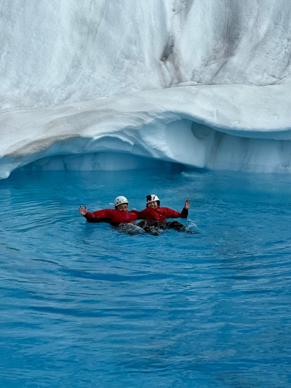 Two swimmers in red drysuits and helmets floating in a bright blue meltwater pool beneath an overhang of glacier ice