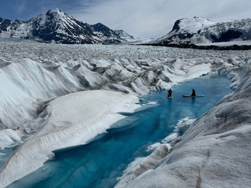 Two paddleboarders on a turquoise meltwater river winding between walls of glacier ice, snow covered peaks beyond