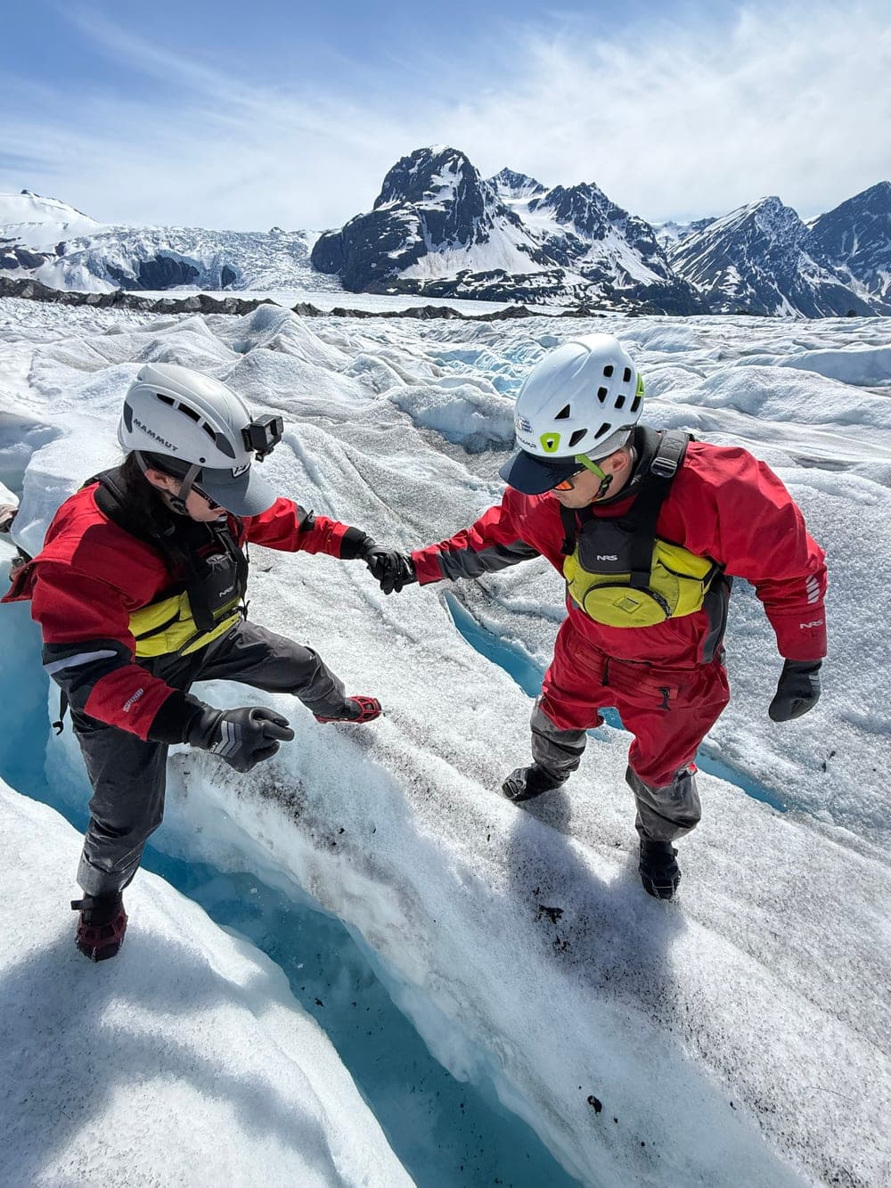 Two people in red drysuits and climbing helmets steadying each other as they step across a crevasse