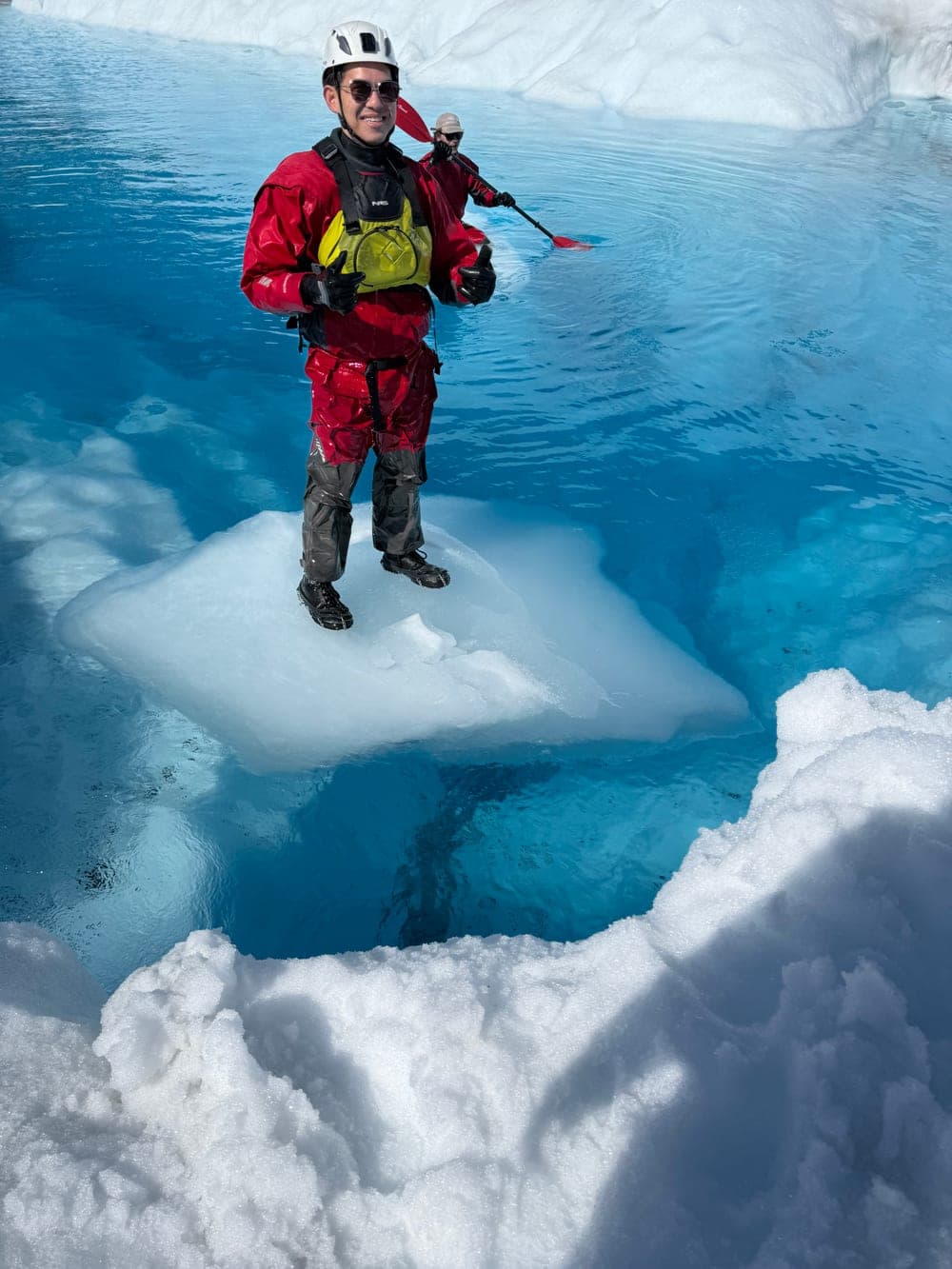 A paddler in a red drysuit balanced on a floating slab of ice in a deep blue meltwater pool, giving a thumbs up