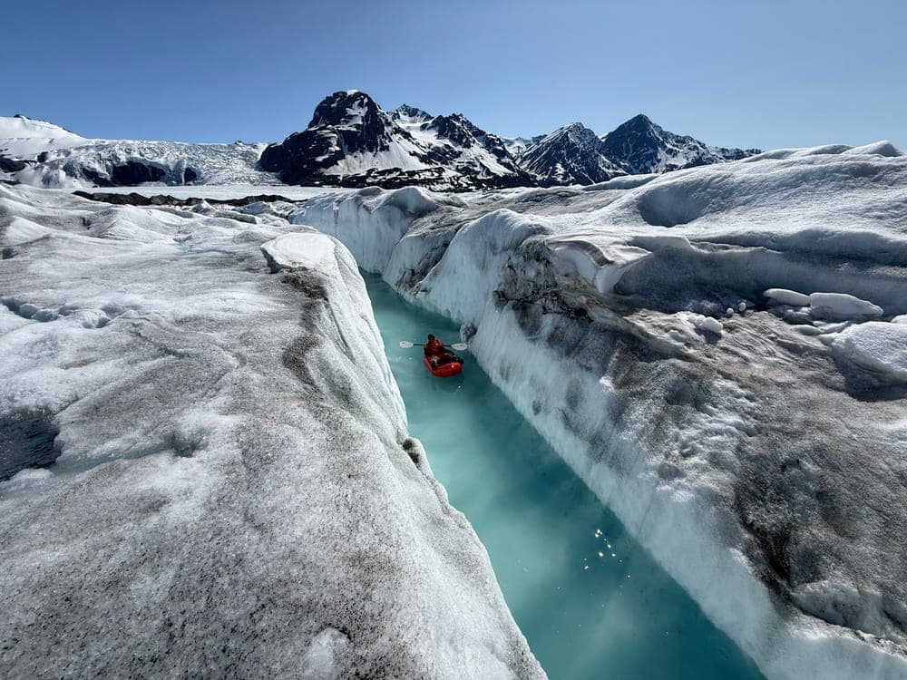 A single red packraft threading a narrow meltwater canyon cut deep into a glacier, jagged peaks on the horizon