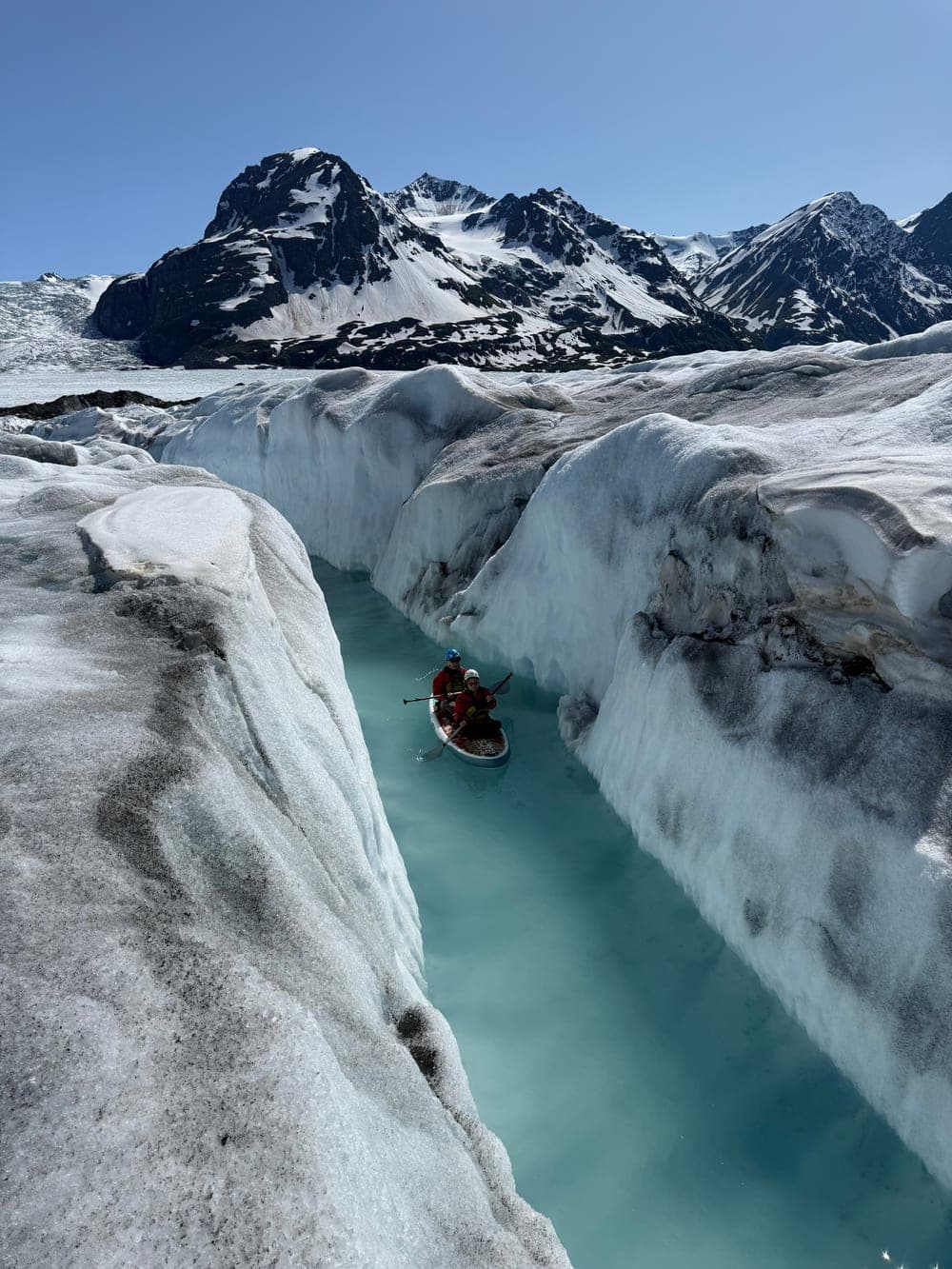 Two paddlers on one board following a narrow meltwater canyon between tall walls of glacier ice