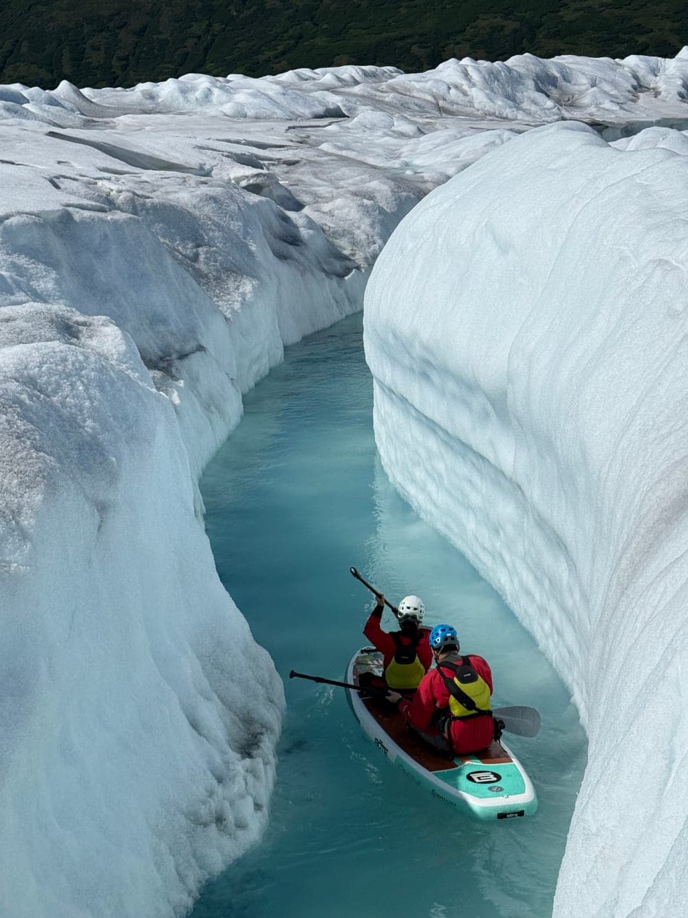 Two paddlers seen from behind, paddling a board down a curving ice canyon on the surface of a glacier