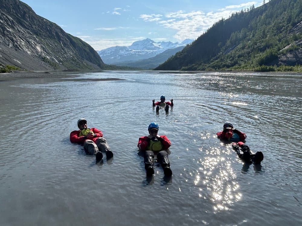 Four people in drysuits floating on their backs down a shallow braided river, snow covered mountains at the head of the valley