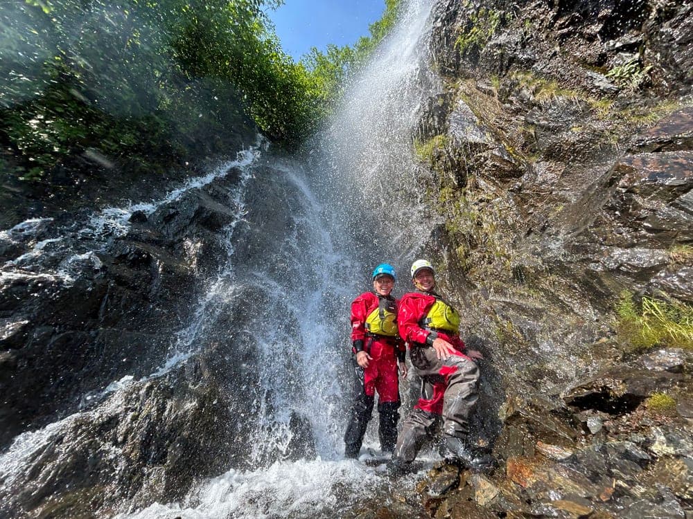 Two people in red drysuits standing and laughing under a waterfall on a mossy rock face