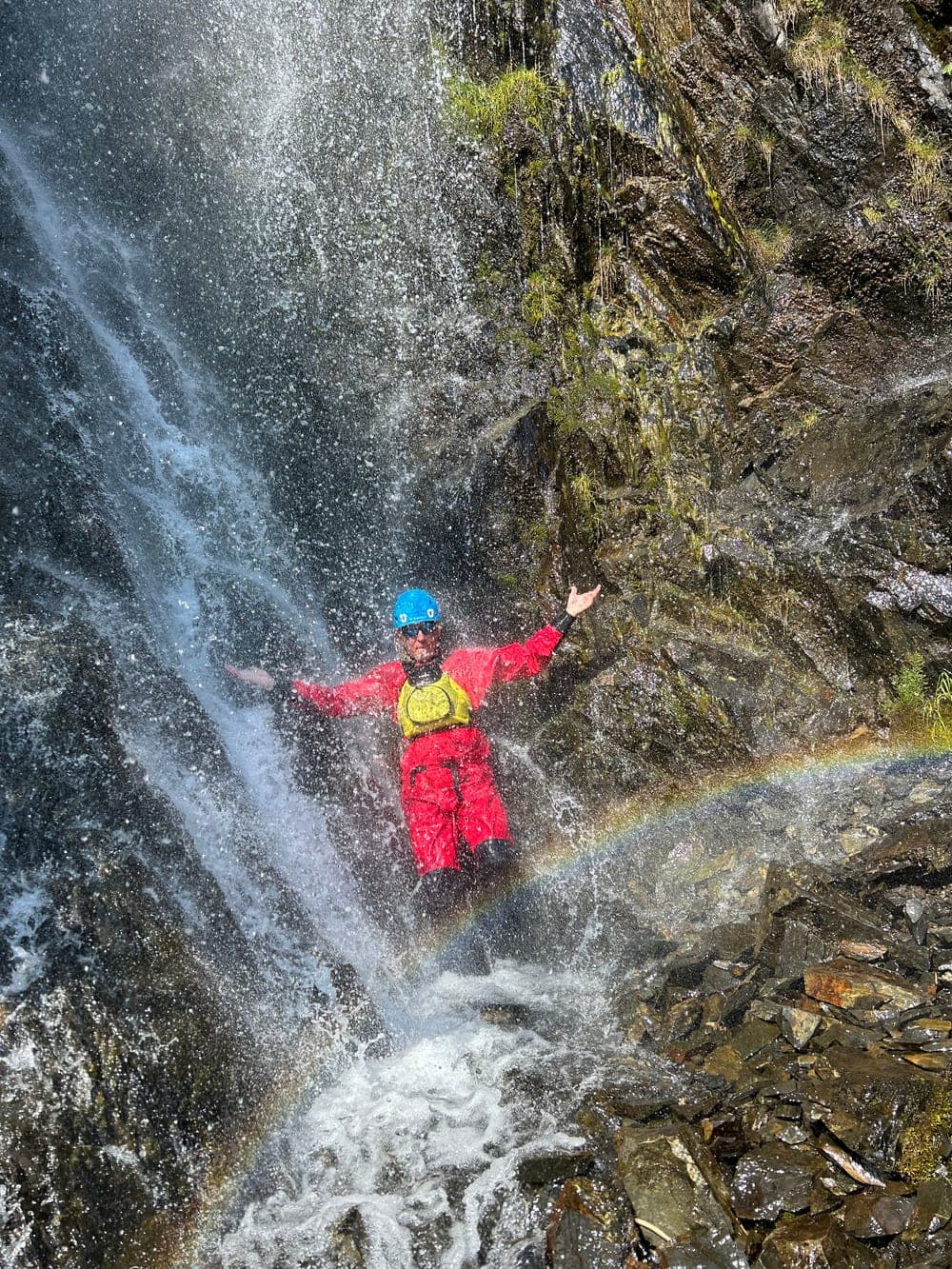 A person in a red drysuit standing under a waterfall with arms outstretched, a rainbow in the spray beside them