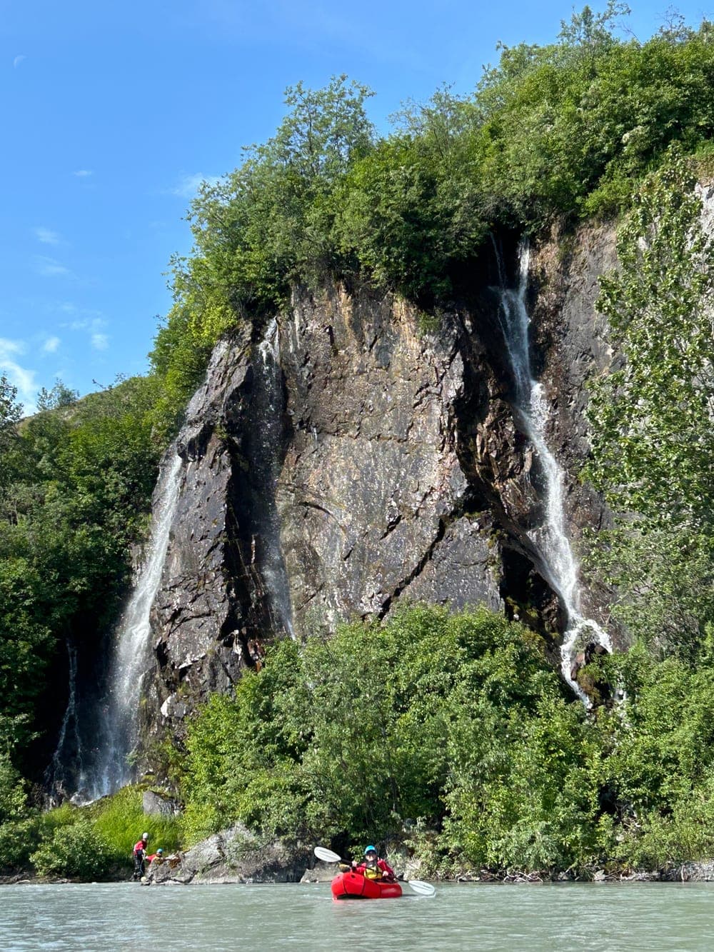 A packrafter on a glacial river below a tall cliff where two waterfalls drop through the trees, two more people waiting on the bank
