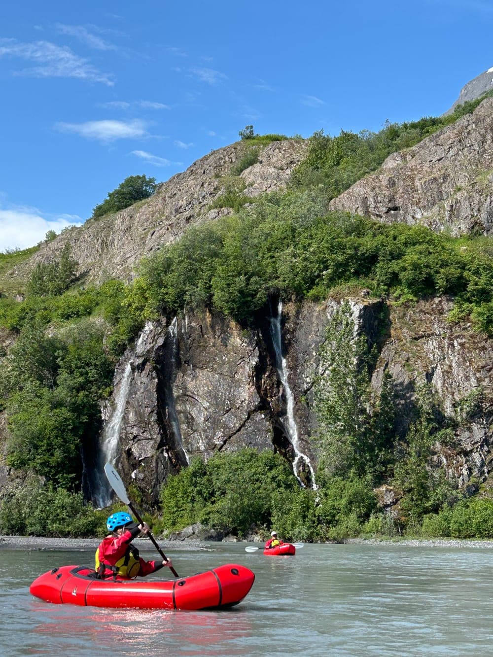 A packrafter paddling a glacial river toward a rock face streaked with twin waterfalls, a second boat further downstream