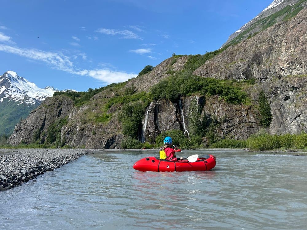 A packrafter crossing a braided glacial river past a gravel bar, waterfalls falling from the cliffs ahead